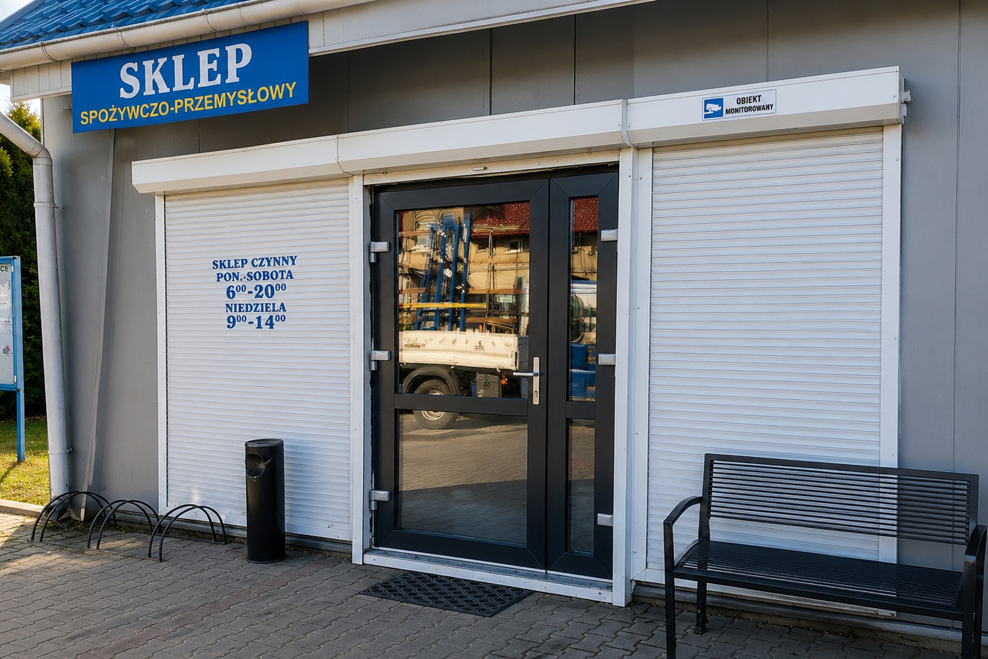 Wejście do sklepu z czarnymi drzwiami aluminiowymi i białymi roletami zewnętrznymi